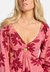 Vrouw die een roze en rode bloemenjurk draagt met opgestroopte mouwen en een ronde uitsnijding bij de borst.