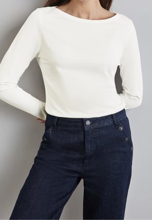 Žena ve jednoduchém bílém tričku s dlouhým rukávem zasunutém do tmavě modrých džínových kalhot s vysokým pasem a bočními knoflíky pózuje u šedé zdi.