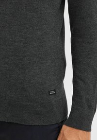 Mørkegrå sweater lavet af blødt strikket materiale. Har en ribbet kant og manchetter, med et lille mærke nederst, der viser "DEDICATED TO DEEP PROTECTION."
