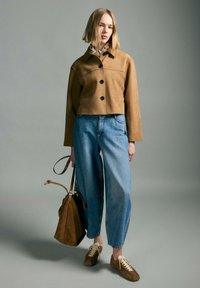 Woman standing in tan jacket, blue high-waisted jeans, brown sneakers, holding brown suede handbag against gray background.
