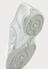 Zapatilla deportiva blanca con parte superior de cuero suave, detalles en plata, suela texturizada con patrón en zigzag y diseño de banda de rodadura circular.