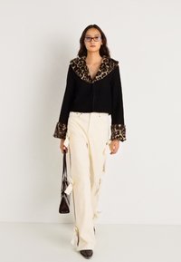 Jaqueta preta cropped com gola e punhos de pelagem sintética de leopardo, combinada com calças largas crème adornadas com laços; modelo segurando uma bolsa.