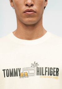 Κοντινό πλάνο ενός ατόμου που φοράει λευκό πουκάμισο με το κείμενο "Tommy Hilfiger Established 1985" και μια εικόνα με δύο κίτρινες καρέκλες και έναν φοίνικα.