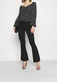 Blusa nera floreale con maniche a sbuffo e scollatura quadrata, abbinata a pantaloni neri a campana e sandali con tacco alto e cinturini.