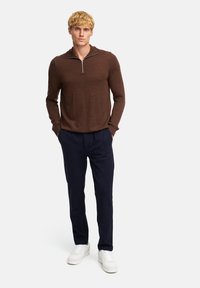 Maglione marrone con zip e maniche lunghe, abbinato a pantaloni blu. Il modello sta in posizione casual con sneakers bianche, le mani nelle tasche.