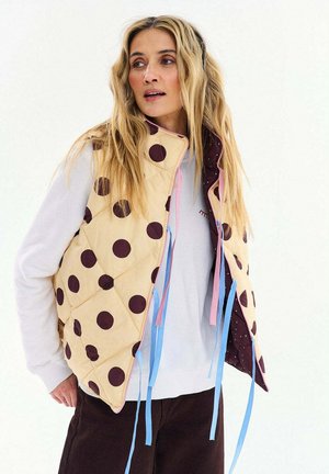 Kobieta ubrana w beżową pikowaną kamizelkę bez rękawów z dużymi brązowymi grochami, na białej koszuli z długim rękawem i ciemnych spodniach, na białym tle.