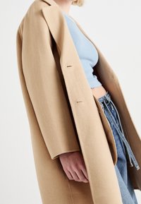 Abrigo clásico de lana camel con corte recto, dos botones frontales y solapas de muesca, combinado con una blusa azul de corte corto y jeans de mezclilla.