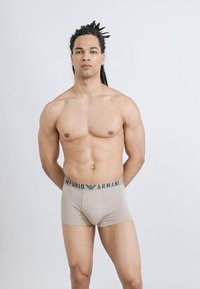 Μυώδης άνδρας με ντρέντλοκς στέκεται αντικρίζοντας μπροστά, φορώντας μπεζ εσώρουχα Emporio Armani μπόξερ, πάνω σε απλό φωτεινό φόντο.