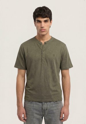 Uomo con capelli corti e scuri che indossa una maglietta henley verde oliva a maniche corte e jeans grigi chiari, in piedi davanti a uno sfondo semplice.