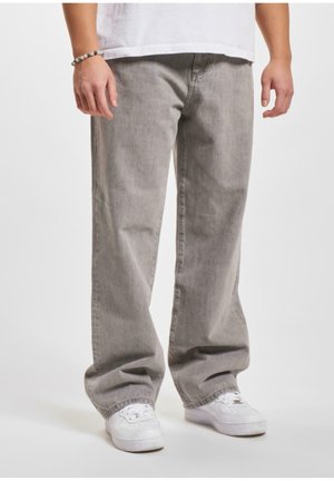 Osoba ubrana w luźne szare jeansy, białe trampki i białą koszulkę, stoi z rękami swobodnie opuszczonymi wzdłuż ciała.