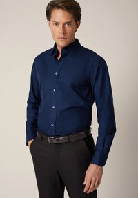 Granatowa koszula z długim rękawem, z usztywnianym kołnierzykiem, zapinana na guziki z kontrastowymi guzikami, zestawiona z ciemnymi spodniami i czarnym paskiem.