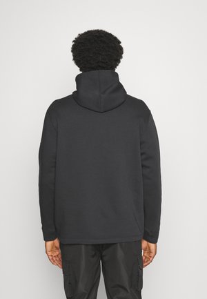 Homme aux cheveux bouclés vu de dos, portant un sweat à capuche noir et un pantalon cargo noir, sur un fond blanc uni.