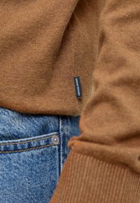 Pruun kudumusega kampsun soonikkoes ja musta brändisildiga, kombineeritud sinise denimiga, millel on hõbedane nupp ja õmbluste detailid.