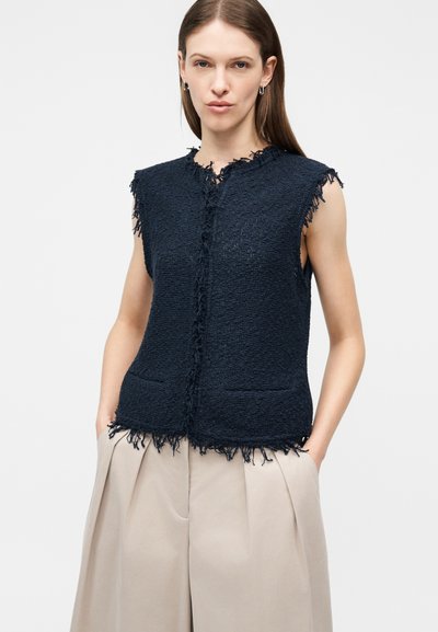 Γυναίκα που φοράει αμάνικο σκούρο μπλε υφασμάτινο γιλέκο με τριχωτές άκρες και μπεζ παντελόνι φαρδιάς γραμμής, στέκεται με τα χέρια στις τσέπες.