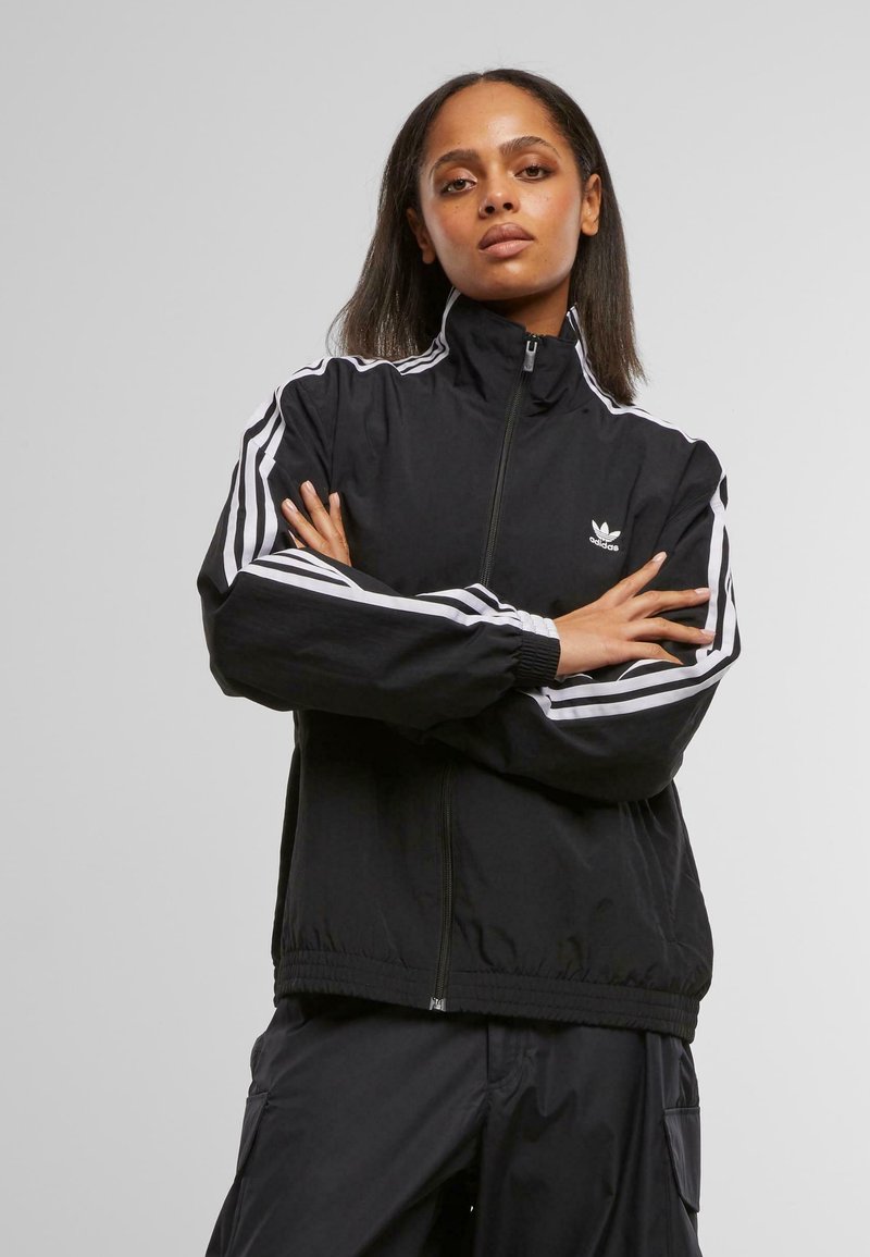 Schwarze Zip-Jacke aus leichtem Stoff. Mit weißen Streifen an den Ärmeln und einem Logo auf der Brust. Weicher Stoff mit elastischen Bündchen und Saum.