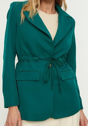 Vrouw draagt een op maat gemaakte teal blazer met een koordje in de taille en klepzakken over een beige top.