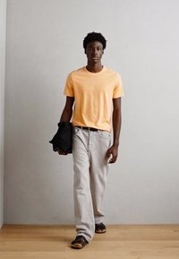 Tricou din bumbac portocalie, blugi din denim gri, jachetă neagră aruncată pe un braț și sandală maro. Simplu, croială lejeră, fără modele vizibile.