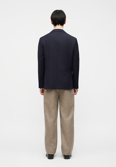 Άνδρας που στέκεται με την πλάτη προς τα εμπρός, φορώντας σκούρο μπλε μπλέιζερ, μπεζ παντελόνι με φαρδύ πόδι και μαύρα παπούτσια, σε απλό λευκό φόντο.