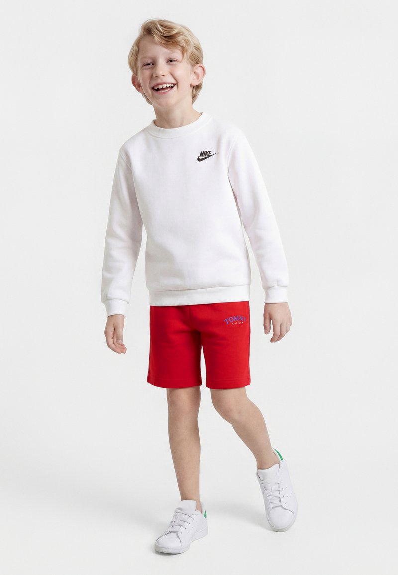 Χαμογελαστός ξανθός αγόρι που περπατά, φοράει λευκό φούτερ Nike, κόκκινα σορτς Tommy Hilfiger και λευκά αθλητικά παπούτσια σε απλό φόντο.