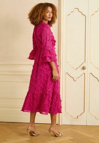Fuksia värvi kleit tekstuursete kaunistustega, volangiliste varrukatega, astmelise heitega ja vöökohal vööga; kombineeritud kuldsete ribadega kontsakingadega.