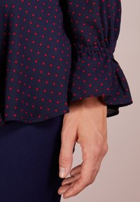 Marinblå blus med röda prickar, med veckad ärmdetalj och mjukt, lätt tyg. Fållen är mjukt rundad.