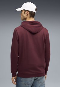 Felpa borgogna con cappuccio e cordino, texture morbida, polsini e orlo ribattuti. Indossata con jeans azzurri chiari e un cappello bianco. Vista da dietro.