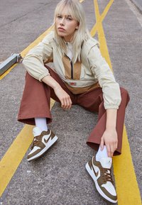 Mladá žena s blond vlasy sedí na silnici mezi žlutými čarami a má na sobě béžovou bundu, hnědé kalhoty, bílé ponožky a bílo-hnědé tenisky Nike.