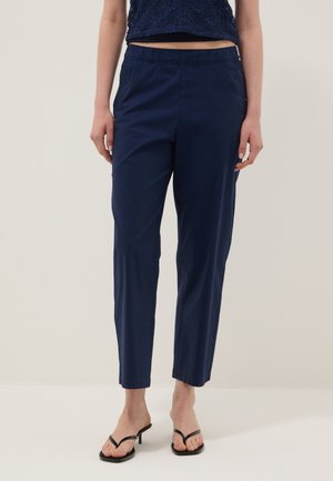 Persoon draagt marineblauwe cropped broek met elastische tailleband en zwarte sandalen met hak, staand tegen een neutrale achtergrond.