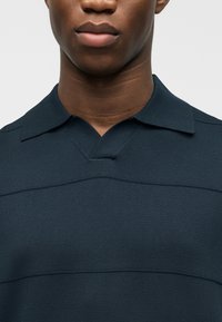 Κοντινό πλάνο του κάτω μέρους του προσώπου και του λαιμού ενός ατόμου που φοράει μια σκούρα μπλε πουλόβερ με γιακά σε σχήμα V και διακριτικές οριζόντιες ραφές.