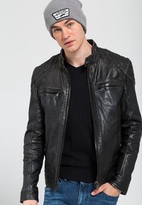 Giacca di pelle nera con chiusura a zip, dettagli trapuntati sulle spalle e due tasche con zip. Indossata sopra un maglione nero a V e abbinata a jeans blu.