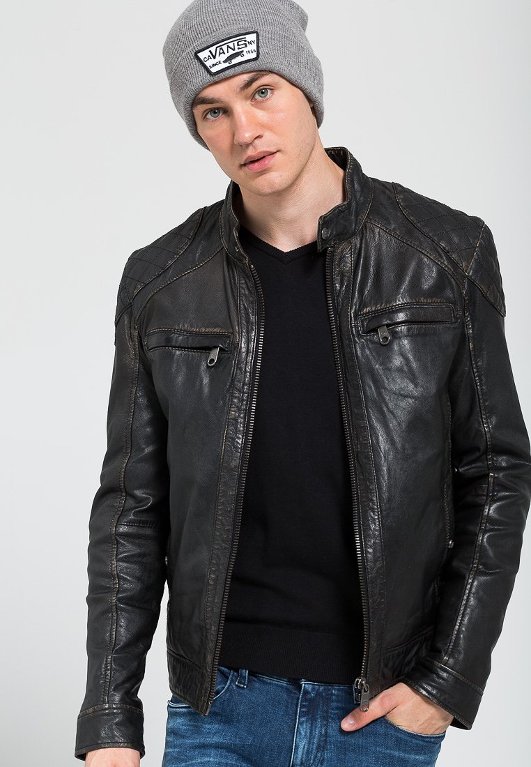 Giacca di pelle nera con chiusura a zip, dettagli trapuntati sulle spalle e due tasche con zip. Indossata sopra un maglione nero a V e abbinata a jeans blu.