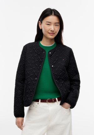 Junge Frau mit langen dunklen Haaren trägt eine schwarze gesteppte Jacke über einem grünen Pullover und weißen Hosen, steht mit einer Hand in der Tasche.