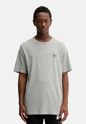 Giovane uomo che indossa una t-shirt grigia Converse All Star a maniche corte e pantaloni neri, in piedi davanti a uno sfondo neutro.