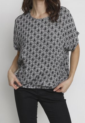 Femme portant un haut ample à manches courtes à motif noir et blanc et un pantalon noir, tenant l'ourlet de la chemise.