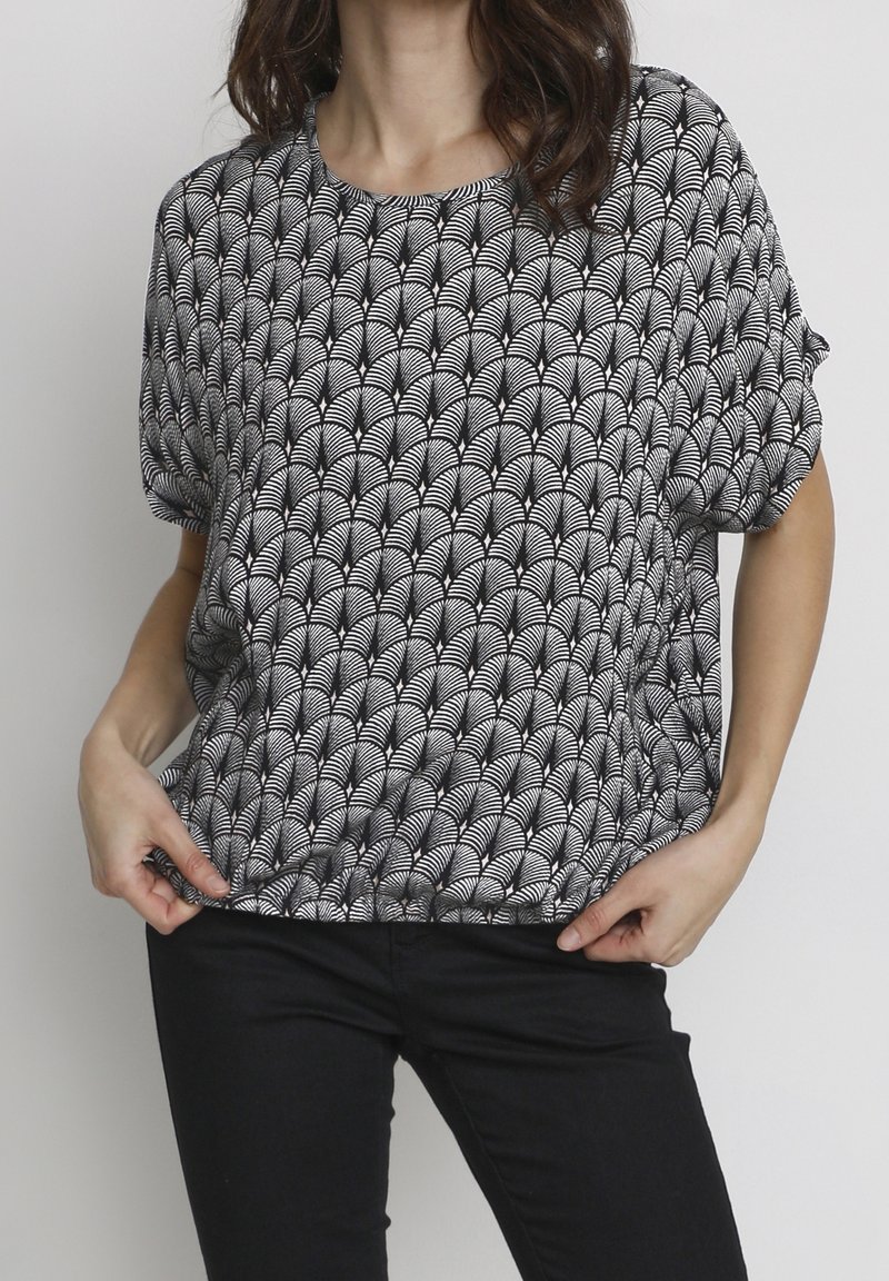 Femme portant un haut ample à manches courtes à motif noir et blanc et un pantalon noir, tenant l'ourlet de la chemise.