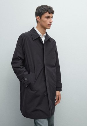 Black, knee-length coat made of smooth fabric, featuring a turn-down collar, front buttons, and side pockets. Worn over a light shirt.