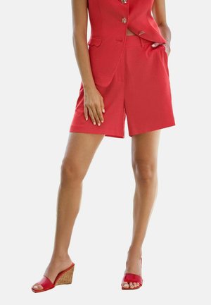 Woman wearing red tailored shorts, matching sleeveless blazer, and red wedge sandals with cork heels standing against a white background.