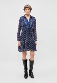 Trumpas rankovėmis mėlynas raštuotas suknelė su apykaklės iškirpte, susiaurinta juosmuo ir plačiai išplatinta sijonu. Juodos iki kelių batai puikiai papildo šį aprangą.