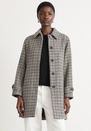 Manteau à carreaux noir et blanc avec un col, des boutons à l'avant et des manches retroussées. Porté sur un haut noir et un pantalon blanc.
