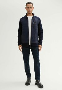 Jachetă navy cu fermoar, cu față matlasată, manșoane ribbate și guler înalt. Purtați-o peste un tricou alb, asociată cu pantaloni închisi la culoare și cizme negre.