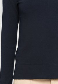 Prim-plan al unei persoane care poartă un pulover tricotat cu mânecă lungă de culoare albastru marin și pantaloni bej, arătând detalii ale trunchiului și brațului.