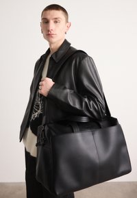 Borsa da viaggio in pelle nera con texture liscia, resistenti manici doppi e un compartimento con zip, tenuta da una persona che indossa una giacca nera.