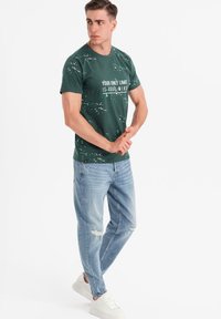 Zelené grafické tričko s bílým textem a designem s rozstřikem, v kombinaci se světle modrými opranými džínami a bílými teniskami.