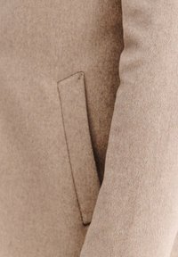 Beżowy wełniany płaszcz o gładkiej fakturze, wyposażony w boczną kieszeń i dopasowany krój. Tkanina ma delikatny połysk i czyste szwy.