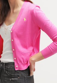 Kvinna i en ljusrosa knäppt cardigan över en vit tanktop med spetsfåll, med handen i fickan på sina mörkgrå jeans.