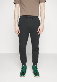 Pantaloni da jogging neri con vita regolabile, caratterizzati da dettagli a pannello e tasche laterali, abbinati a sneakers alte verdi e nere.