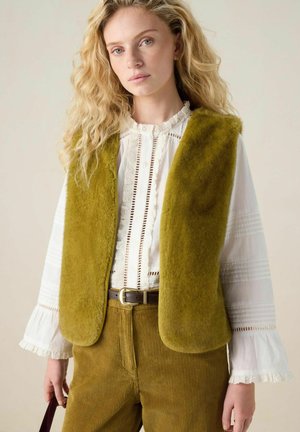 Olive green faux fur vest layered over a white blouse with lace trim, paired with textured mustard corduroy pants. Brown belt visible.