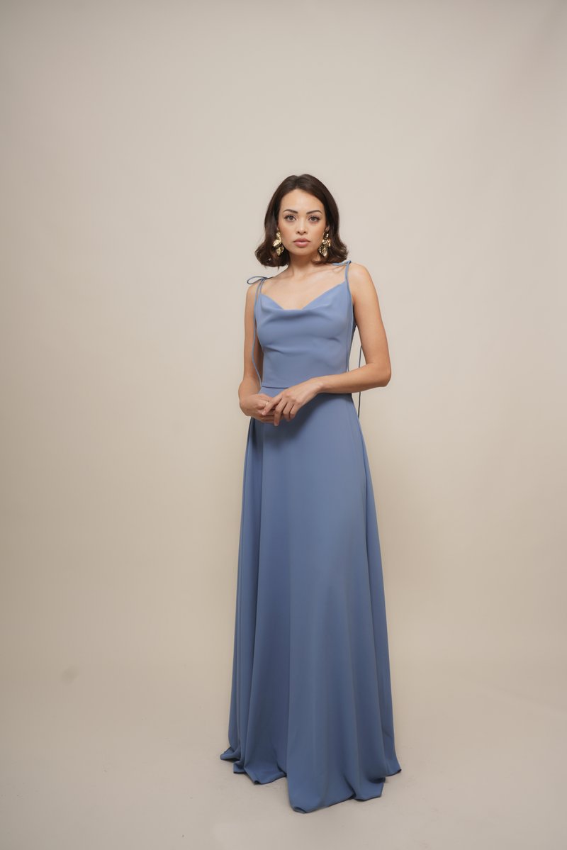 Woman in a blue floor-length gown with thin straps, standing against a plain beige background, wearing large gold earrings.