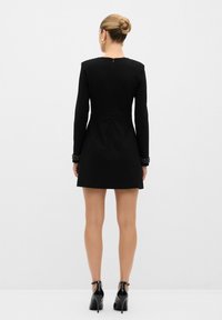 Robe noire à manches longues et silhouette ajustée. Finitions ornées aux poignets et fermeture éclair à l'arrière. Portée avec des chaussures à talons hauts noires.