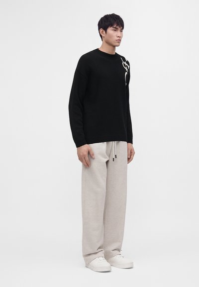 Jeune homme debout portant un pull noir avec un motif blanc, un pantalon beige clair à cordon et des baskets blanches sur un fond uni.